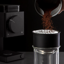 Close-up of coffee grounds being poured from a container into a black and silver coffee dripper placed on a glass. The Fellow Ode Coffee Grinder Gen 2.0 in Matte Black, featuring a large dial for adjusting grind settings, is visible in the background on the left side. The scene is set on a dark surface with a dark background, ready to brew coffee.