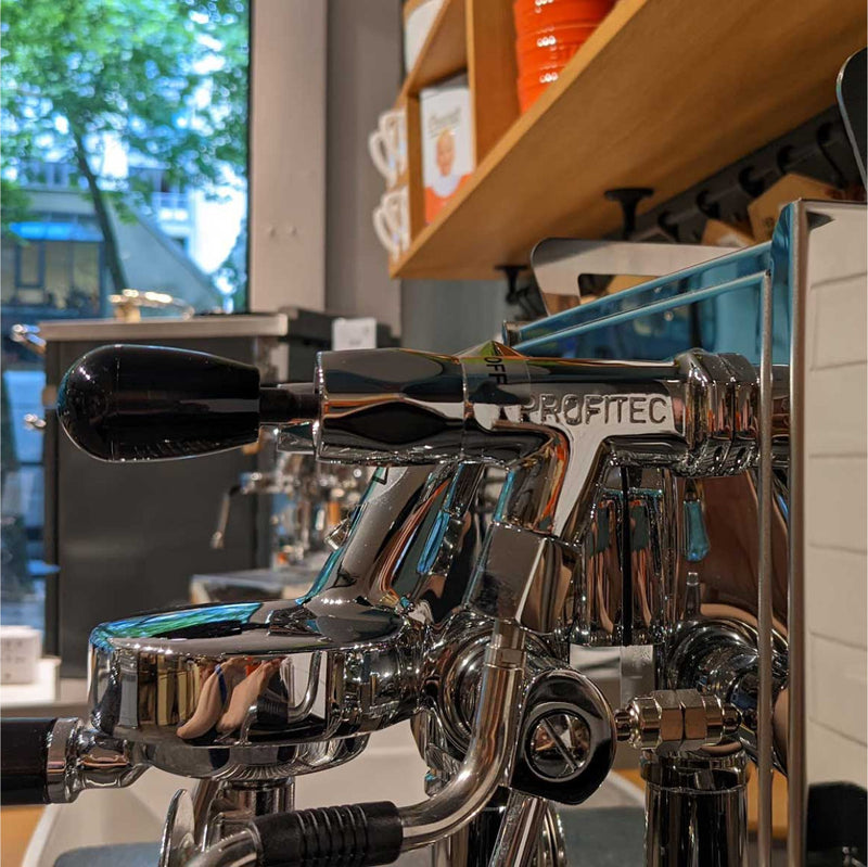 Close-up of a shiny, chrome Profitec Pro 500 Heat Exchanger & Quick Steam Espresso Machine with E61 Group Head. The handle is prominently visible in a cafe setting, with shelves of mugs and coffee-related items in the background, beneath large windows letting in natural light. Its stainless steel boiler adds to the sleek design.