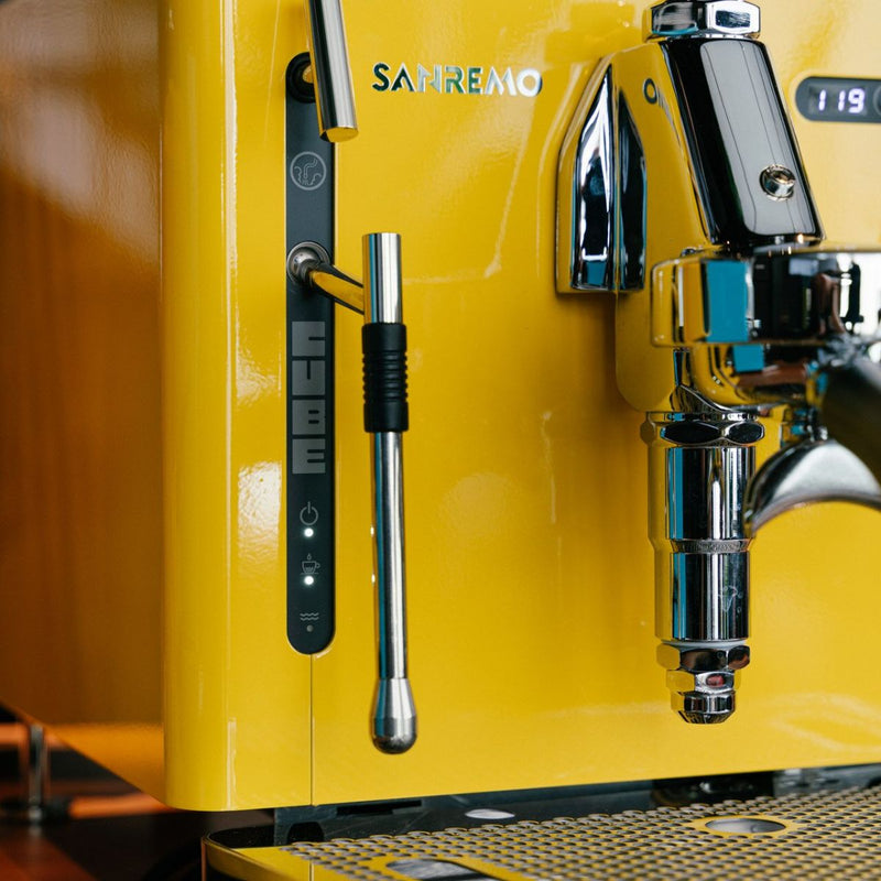 Close-up of a yellow Sanremo Cube R Heat Exchanger Espresso Machine E61 Group Head featuring a metal steam wand, a lever for adjusting settings, and a digital display showing "119." The brand name "Sanremo" is visible on the machine, which also boasts Wi-Fi connectivity.