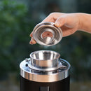 A hand is holding a metallic lid above a DF64 Gen 2 Single Dose Coffee Grinder with DLC Burrs (Black) from DF Grinders. The grinder’s wide opening and adjustment dial are visible, showcasing its precision. The background is blurred, featuring natural green colors. Equipped with 64mm DLC burrs, this open box, unused grinder ensures zero retention grinding for a consistent coffee experience.