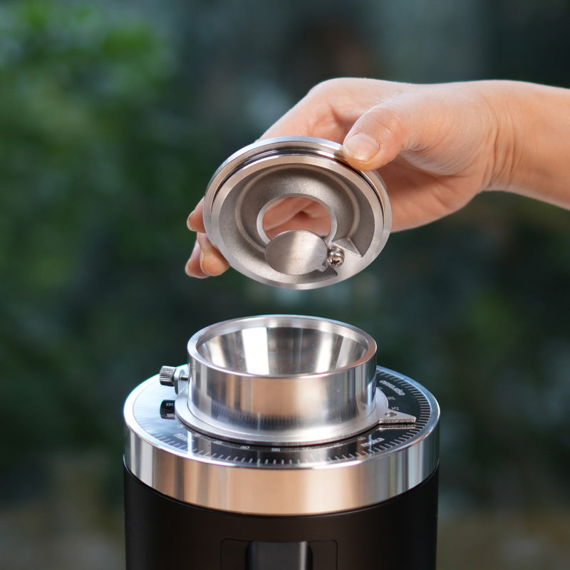 A hand is holding a metallic lid above a DF64 Gen 2 Single Dose Coffee Grinder with DLC Burrs (Black) from DF Grinders. The grinder’s wide opening and adjustment dial are visible, showcasing its precision. The background is blurred, featuring natural green colors. Equipped with 64mm DLC burrs, this open box, unused grinder ensures zero retention grinding for a consistent coffee experience.