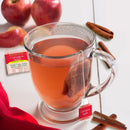 A clear glass mug filled with caffeine-free Bigelow Cinnamon Apple Tea sits on a table. Nearby, there are apples, cinnamon sticks, and a wrapper from the Bigelow Cinnamon Apple Tea Bags. A red cloth is partially visible in the bottom left corner.