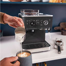 Close-up of hands preparing a cappuccino with a milk frother. One hand holds a pitcher of frothed milk being poured into a cup with latte art, while the other adjusts the KitchenAid Semi Automatic Espresso Machine with Burr Grinder KES6551BL (Cast Iron Black) in the background.