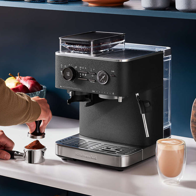 A person prepares the KitchenAid Semi Automatic Espresso Machine with Burr Grinder KES6551BL (Cast Iron Black) by placing ground coffee into the portafilter. The sleek, black espresso machine features a clear water reservoir and has a freshly made cup of coffee sitting next to it on the counter. In the background, fruits and utensils are visible, hinting at a bustling kitchen scene.