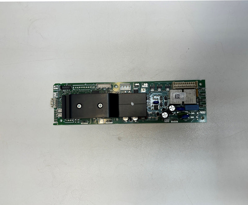 The DeLonghi Parts: Power Card 5213229121 features a green circuit board adorned with various electronic components such as capacitors, resistors, a black rectangular block, and a modular chip section. This circuit board is positioned on a plain light-colored surface.