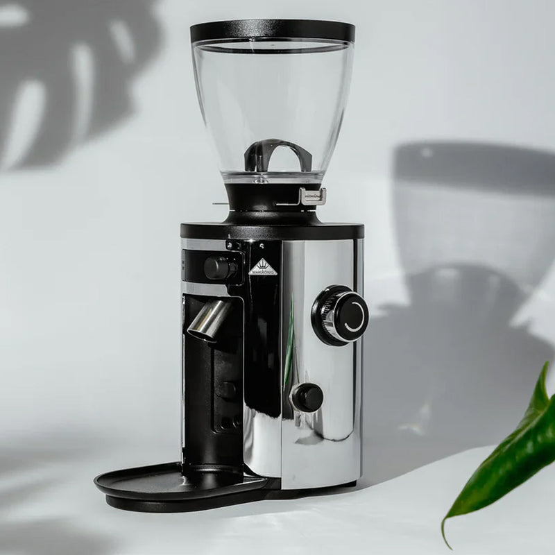 The MAHLKÖNIG X54 Allround Home Grinder (Chrome), featuring a transparent bean hopper on top, a glossy metallic body, control knobs on the side, and a portafilter holder beneath the grinder spout, is set on a minimalistic light-colored surface with some plant shadows surrounding it.