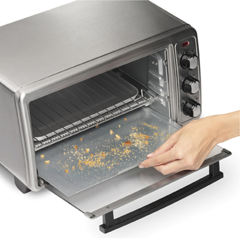 A person is pulling out a crumb tray from the Hamilton Beach Sure-Crisp Air Fryer Toaster Oven 31413. The versatile appliance, with its three knobs and red indicator light on the side panel, also offers air frying capabilities. The tray has some crumb remnants on it.