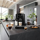 A modern kitchen showcases the DeLonghi Magnifica Start Super Automatic Espresso Machine with LatteCrema System ECAM22080B in black, featuring integrated grinding technology on a dark countertop. The setup is adorned with customizable coffee drinks in glass cups. Nearby, a plate of pastries completes the scene, set against a backdrop of plants and large windows offering a picturesque view.