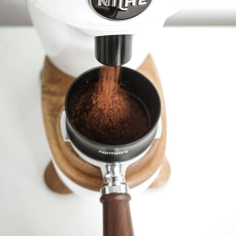 A close-up of ground coffee being dispensed into a portafilter from a white coffee grinder equipped with the Normcore 58mm Dosing Funnel by Normcore. The grinder is positioned on a wooden base, and the handle of the portafilter is made of dark wood. The finely ground coffee appears ready for brewing.