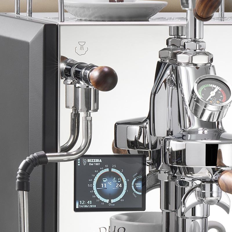 Close-up of a modern Bezzera DUO MN Dual Boiler Espresso Machine w/ Flow Control (White), highlighting its E61 group head with a digital control panel displaying various settings and a manometer indicating pressure. The espresso machine from Bezzera features polished chrome components and wooden handles.