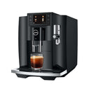 The Jura E8 Piano Black Automatic Coffee Machine 15648 (Latest Version) is a sleek, black coffee machine with a digital display screen showcasing drink options. This barista-quality machine features a built-in milk frother and an espresso shot being dispensed into a glass cup on the tray beneath.