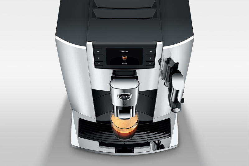 A top view of the Jura E8 Chrome Automatic Coffee Machine 15646 (Latest Version) effortlessly dispensing barista-quality coffee into a small, transparent glass cup. The machine boasts a sleek, metallic chrome design with a digital display panel indicating "Coffee." A steam wand is conveniently located on the right side of the machine.