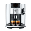 A sleek, modern Jura E8 Chrome Automatic Coffee Machine 15646 (Latest Version) with a digital display showing different beverage options. Two glass cups filled with barista-quality espresso are placed under the dual spouts. The machine has a metallic finish and a side-mounted milk frother.