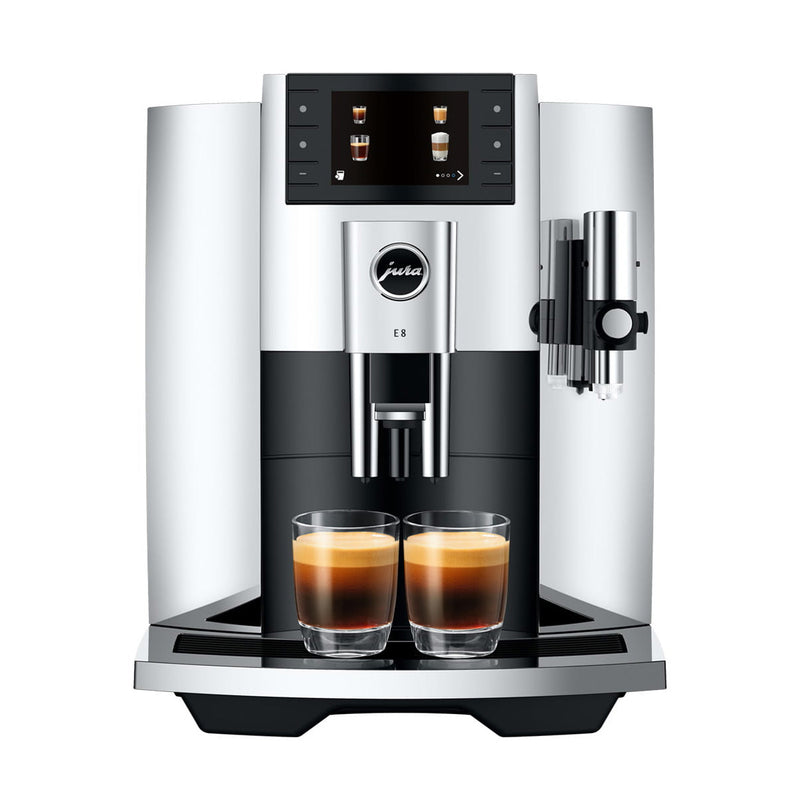 A sleek, modern Jura E8 Chrome Automatic Coffee Machine 15646 (Latest Version) with a digital display showing different beverage options. Two glass cups filled with barista-quality espresso are placed under the dual spouts. The machine has a metallic finish and a side-mounted milk frother.