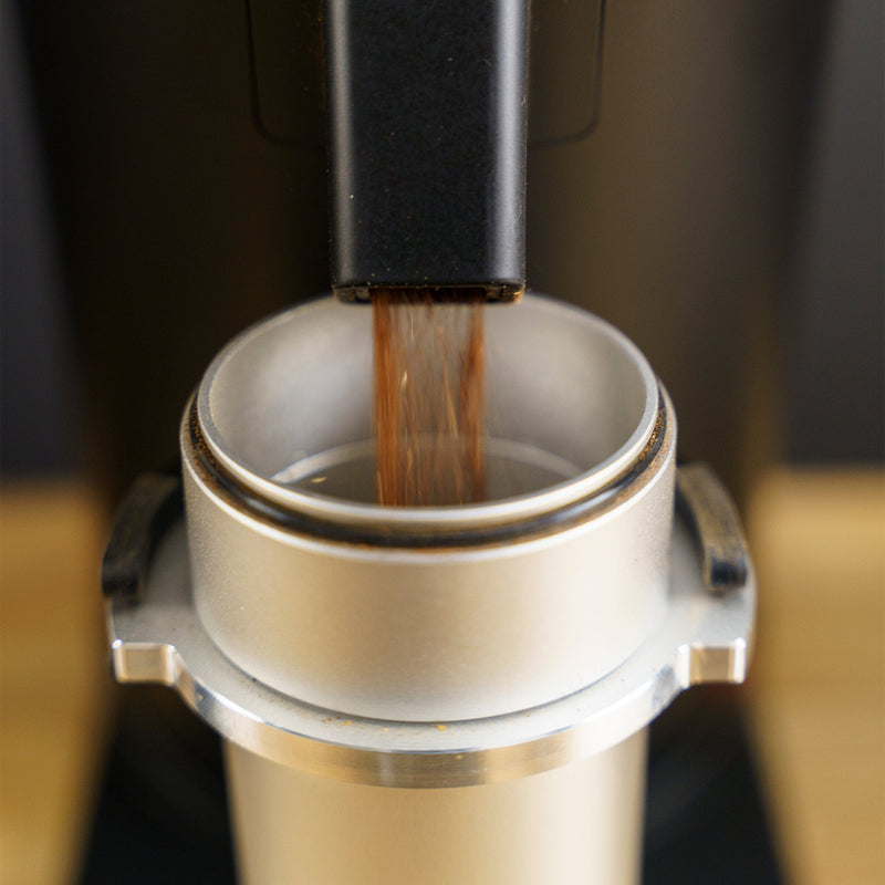 Close-up of freshly ground coffee being dispensed from a DF64 Gen 2 Single Dose Coffee Grinder With DLC Burrs (Black) by DF Grinders into a metallic portafilter. The portafilter is seated beneath the grinder’s chute, ready to be tamped and used for brewing espresso. The background is blurred, emphasizing the zero retention grinding process.