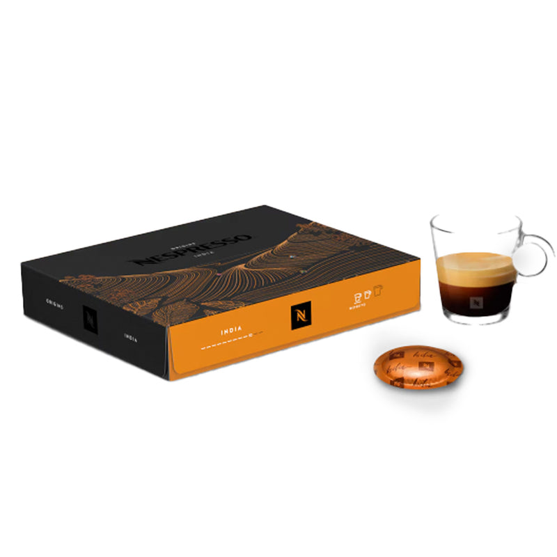 A box of Nespresso Ristretto Origin India Professional Capsules, featuring intricate designs, is placed next to a glass cup containing a spicy blend of espresso. One Arabica Robusta blend capsule is lying beside the box.