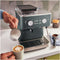 A person pours steamed milk from a pitcher into a cup of coffee with latte art, in front of a KitchenAid Semi Automatic Espresso Machine with Burr Grinder KES6551JP (Juniper) on a kitchen countertop. The machine features buttons, dials, and a steam wand, with coffee beans visible in the burr grinder container on top.