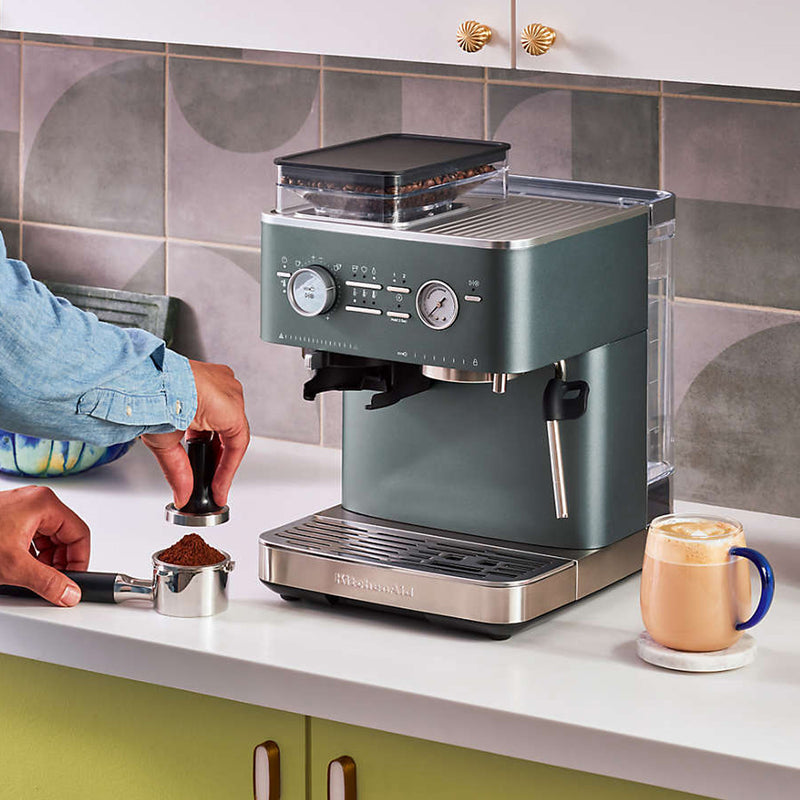 A person in a blue shirt is tamping ground coffee beside the KitchenAid Semi Automatic Espresso Machine with Burr Grinder KES6551JP (Juniper) on a kitchen counter. A cup of prepared coffee sits to the right of the machine. The background features a tile backsplash and part of a cabinet with green drawers.