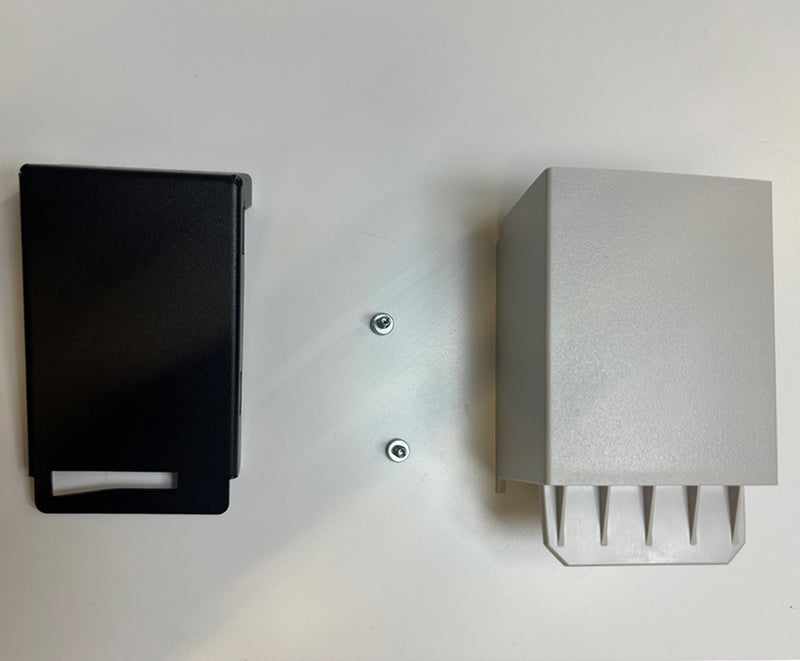 Two rectangular plastic enclosures are mounted on a light gray surface. The left enclosure is black and features an open-bottom design similar to the Keurig VKI Eccellenza Touch LOCK ASM HOPPER (76-39246). The right enclosure is gray with a solid structure and has four protruding slots at the bottom. Two screws are visible between them.