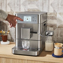 On a wooden counter, there is a KitchenAid KF7 Fully Automatic Espresso Machine KES8557SX in brushed stainless steel. A person is touching the control panel on the machine. Beside the espresso machine, there's a glass of milk, a container of milk connected to the machine for automatic milk frothing, and a cup of coffee with froth.