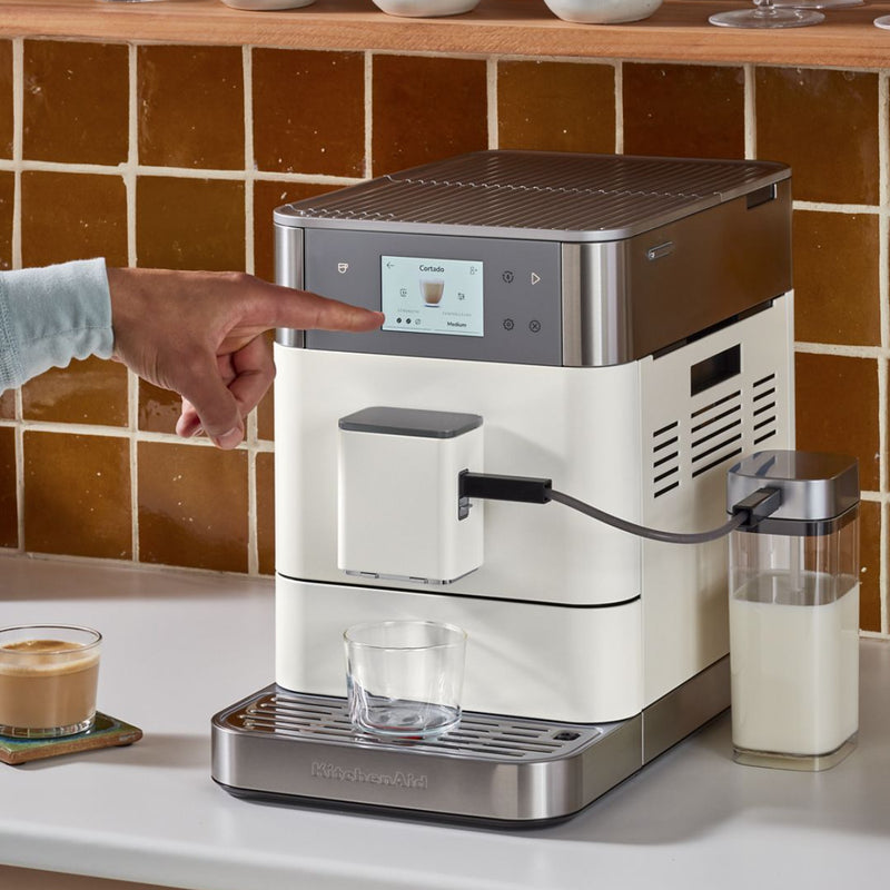 A person is selecting a "Cortado" on the sleek KitchenAid KF7 Super Automatic Espresso Machine KES8557PL, which features a digital screen. A glass waits below the spout as the automatic milk frothing begins. Nearby, a latte sits on the countertop against the backdrop of brown tiles.