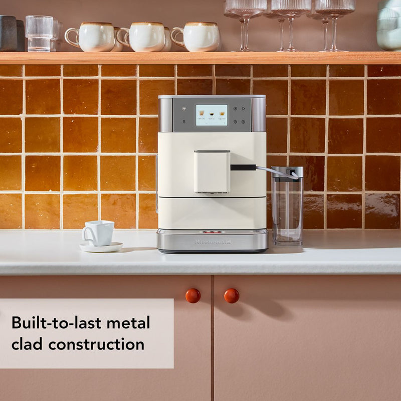 A KitchenAid KF7 Super Automatic Espresso Machine KES8557PL in Porcelain graces a kitchen counter against a retro tile backsplash and pastel cabinets. Cups are neatly arranged on the shelf above. The text reads, "Built-to-last metal clad construction featuring personalized drinks and automatic milk frothing.