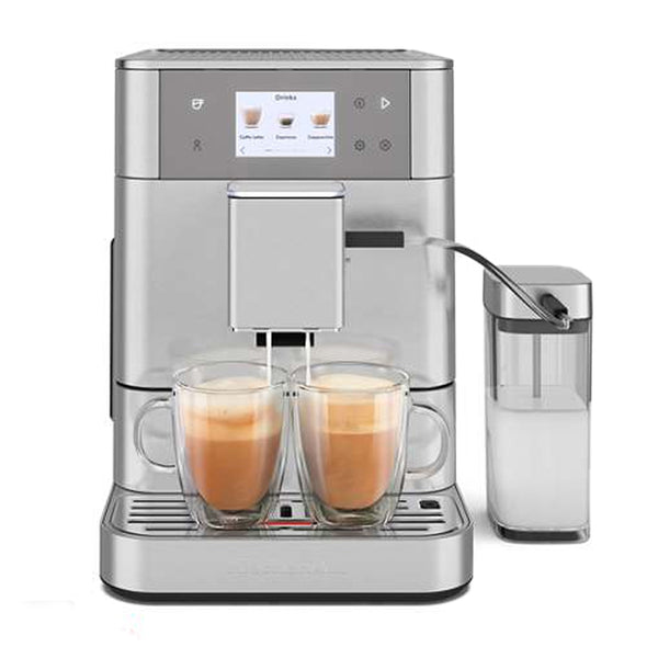 The KitchenAid KF7 Fully Automatic Espresso Machine KES8557SX, in brushed stainless steel, boasts a digital display and the ability to brew two cups of coffee simultaneously. It features automatic milk frothing with a transparent milk container on the right side and includes two glass coffee cups placed on the drip tray for personalized drinks.