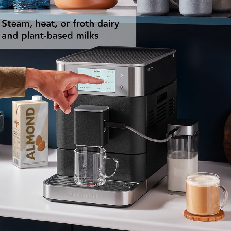 A person is using a KitchenAid KF8 Fully Automatic Espresso Machine (Cast Iron Black) KES8558BK with a touchscreen interface to make a barista-quality beverage. An almond milk carton is next to the machine. The text on the image reads, "Steam, heat, or froth dairy and plant-based milks.