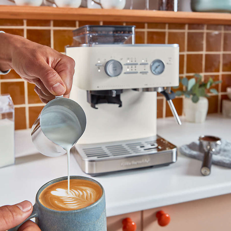 A person pours steamed milk with latte art into a cup of coffee, while the elegant KitchenAid Semi Automatic Espresso Machine with Burr Grinder KES6551PL, in a cream-colored porcelain finish and featuring smart dosing technology, sits gracefully on the kitchen counter against the tiled backsplash.