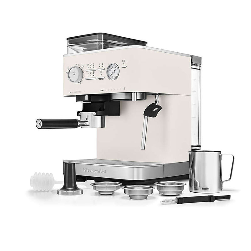 The KitchenAid Semi Automatic Espresso Machine with Burr Grinder KES6551PL, in a cream-colored porcelain finish, comes equipped with a steam wand and cup warmer. It includes multiple metal filter baskets, a tamper, milk frother jug, and cleaning tool. The machine boasts smart dosing technology on its control panel, featuring dials and buttons for precise operation.