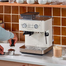 A person is preparing to make coffee using the KitchenAid Semi Automatic Espresso Machine with Burr Grinder KES6551PL, featuring smart dosing technology, on a kitchen counter. The porcelain machine with silver accents sits beside a freshly made cup of coffee topped with milk froth, while a brown tile backsplash adds warmth to the scene.