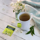 A steaming mug of Bigelow Jasmine Green Tea sits on a white wooden table alongside its tea bag and packaging. A small floral arrangement in a glass jar and a few green tea leaves add to the scene, with a soft blue cloth in the background, enhancing the delicate floral aroma that fills the air.