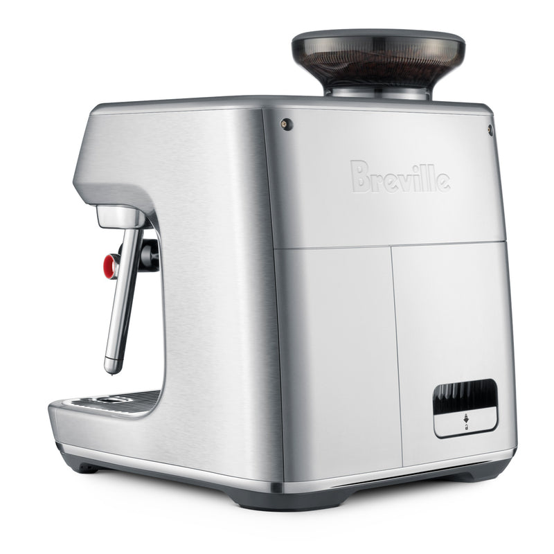 A sleek, brushed stainless steel Breville Oracle Jet Espresso Machine (BES985BSS) with a built-in grinder sits angled slightly to the right. The machine features a steam wand on the left side and a control knob on the side, combining commercial performance with advanced automation. The Breville logo is prominently displayed on the front.