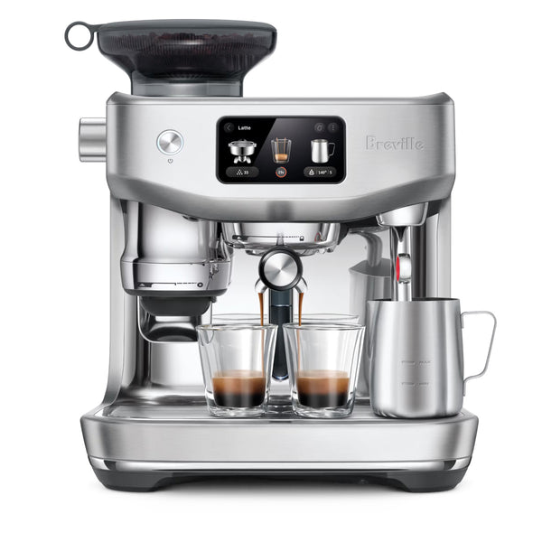 The Breville Oracle Jet Espresso Machine BES985BSS (Brushed Stainless Steel) by Breville offers advanced automation for commercial performance with its sleek design and digital display. Two shot glasses are being filled with espresso, a frothing pitcher rests on the steam wand, and a grinder is integrated on top. The machine displays settings and icons for various coffee options.