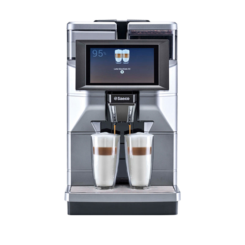 A sleek, silver Saeco Magic M2+ Commercial Coffee Machine with a digital display shows two cups of latte macchiato. Below the display, two nearly identical customizable beverages are being dispensed simultaneously into tall, clear glass cups placed on the coffee machine's tray.