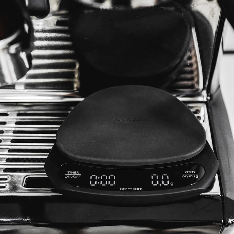 A Normcore Ultra Thin Coffee Scale, finished in sleek black, rests on a gleaming metallic espresso machine drip tray. The minimalist-designed scale displays zeros for both weight and timer on its digital screen. The name "Normcore" is prominently featured, highlighting its high precision weighing accuracy.