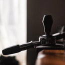 Close-up of a portafilter inserted into an espresso machine. The handle of the portafilter extends to the left, while the Normcore Spring Loaded Tamper V4 (58mm) is positioned vertically on top. The background is softly lit, contrasting with the sharp focus on the coffee-making equipment from Normcore.