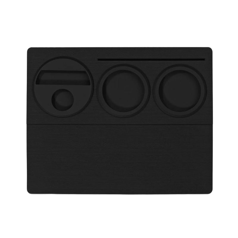 The Normcore Espresso Tamping Station V2, a black desktop organizer made from durable silicone rubber, features multiple compartments of various shapes and sizes including a rectangular slot, two circular compartments, and a small circular hole. This minimalist piece is perfect for organizing your espresso accessories.