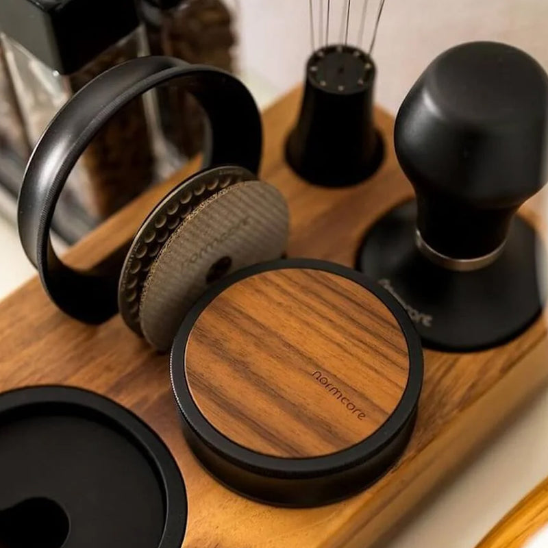 The Normcore Narrow Espresso Tamping Station - Walnut from Normcore holds various coffee-making tools, including a portafilter handler, a coffee tamper, and a distribution tool. Perfect for any coffee bar or Espresso Tamping Station, the tray’s minimalist design highlights wooden and black metal elements and accommodates 54mm and 58mm espresso accessories.