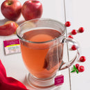 A clear glass cup filled with Bigelow Cranberry Apple Tea, an all-natural, caffeine-free beverage, with a teabag steeping inside. The cup is placed on a white surface alongside scattered cranberries, apples, and a red cloth. A box of Bigelow Cranberry Apple Tea Bags is in the background.