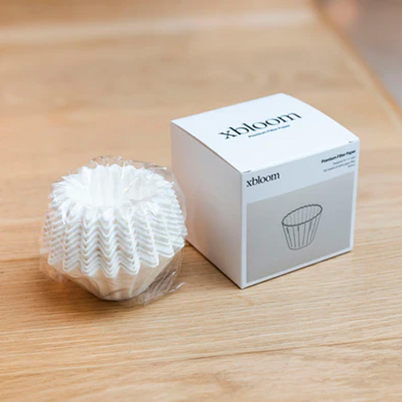 A pack of xBloom Paper Filters (Pack of 100) wrapped in plastic is placed next to a minimalist white box labeled "xBloom Premium Filter Paper" on a wooden surface, showcasing customized premium wave paper filters for your Omni Dripper.