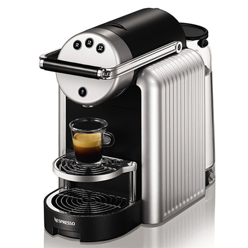 A Nespresso Professional Zenius Machine, featuring a sleek silver and black exterior, is displayed with a small glass espresso cup partially filled with espresso positioned on the drip tray. The machine has a cylindrical design and includes three buttons at the top front for making different types of coffee, making it ideal for self-serve spaces.