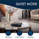 A person sits on a couch holding a book and a glass bottle. On the round table in front of them is a glass of water and a DeLonghi Pinguino Arctic Whisper Portable Air Conditioner, Up To 700 sq. ft. EX390LVYN (Black) displaying "Quiet Mode." The text "IDEAL FOR" is below with icons labeled "sleeping," "watching TV," and "reading," highlighting its Cool Surround Technology.