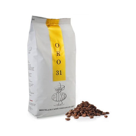 A white and yellow bag of Caffe Mike Oro 31 Espresso, containing 1kg (2.2lbs) of whole bean coffee, stands upright next to a small pile of coffee beans. The bag features Italian text and a logo with the letters "MKF." The packaging is simple and clean, suggesting a premium dark roast coffee product produced through small batch roasting.