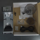 Top view of an open box containing a Baratza Sette 30 AP (All Purpose) Conical Burr Coffee Grinder (2562) with 31 stepped grind adjustments. Inside, you can also find a cleaning brush, manual, two clear plastic containers, a metal tool, small packet, rubber piece, and power cord all arranged neatly.