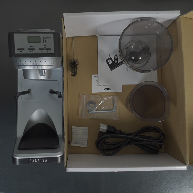 Top view of an open box containing a Baratza Sette 30 AP (All Purpose) Conical Burr Coffee Grinder (2562) with 31 stepped grind adjustments. Inside, you can also find a cleaning brush, manual, two clear plastic containers, a metal tool, small packet, rubber piece, and power cord all arranged neatly.