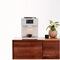 A Terra Kaffe TK-01 Super Automatic Espresso, Cappuccino, & Latte Machine (White) - OPEN BOX (3397) sits on a wooden surface, with a small cup positioned under the dispenser. The background features a potted plant with green leaves and a white wall. The machine displays multiple buttons and a small, lit space where the coffee is dispensed, ideal for any cozy kitchen setup.